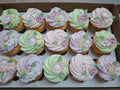 A box of decorated cupcakes with pastel pink, lavender, and mint green frosting, topped with 3D flowers and green leaves.