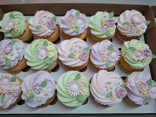 A box of decorated cupcakes with pastel pink, lavender, and mint green frosting, topped with 3D flowers and green leaves.